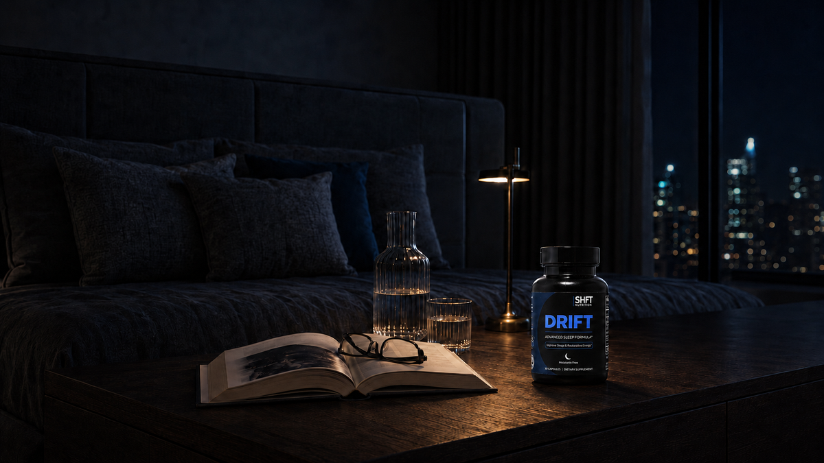 A bottle of Drift sleep supplement placed on a dark wooden table in a dimly lit bedroom at night, with an open book, glasses, a glass carafe, and a lit lamp nearby.
