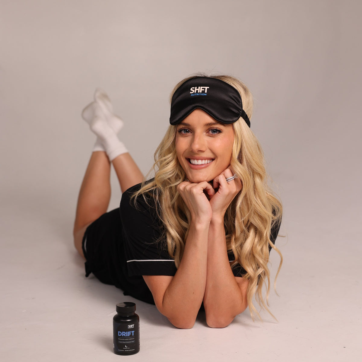 Lindsay Brewer smiling with an SHFT sleep mask pushed up on her head, lying next to a bottle of DRIFT