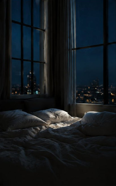 A dimly lit bed with rumpled white bedding in a dark bedroom, with tall windows showing a nighttime city skyline outside.