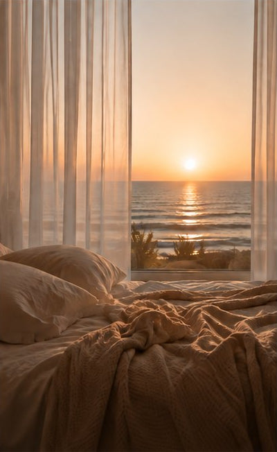 A cozy bed with rumpled beige bedding and pillows, framed by sheer white curtains, looking out onto an ocean sunset at the beach.