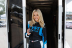 Lindsay Brewer in a black and blue racing suit stands in the doorway of a trailer, holding a bottle of DRIFT. The suit features the text 'SHFT NUTRITION'.