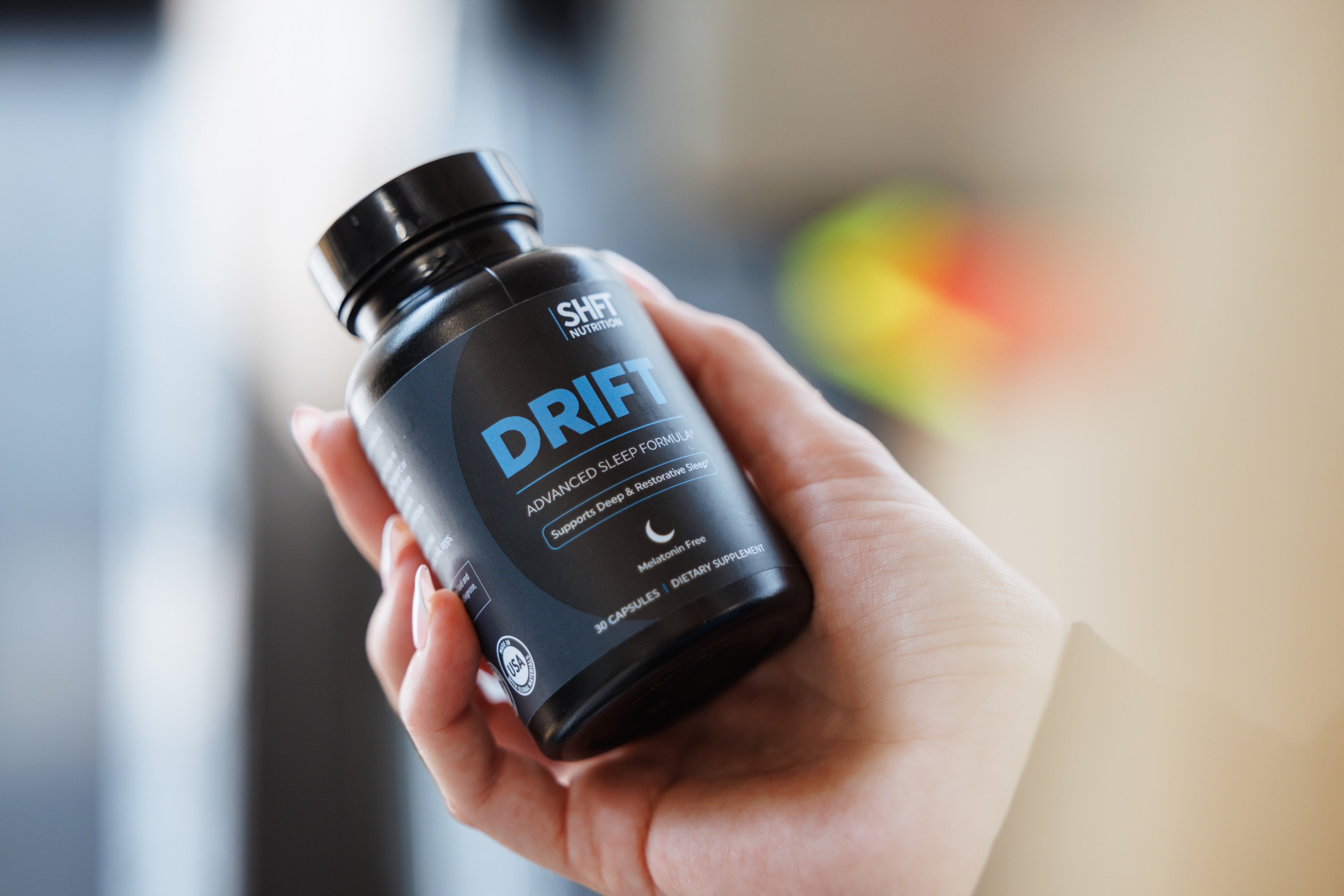 A hand holding a black bottle of SHIFT LABS DRIFT Advanced Sleep Formula capsules, with a blurred indoor background.